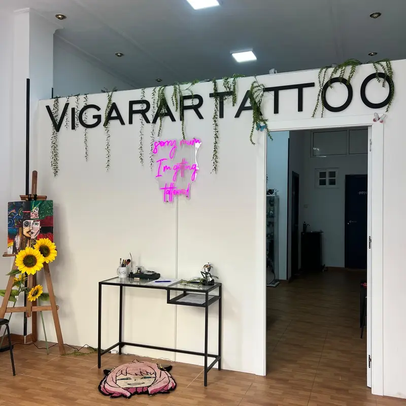 Entrada del estudio Vigarartattoo con el rótulo grande en la pared, plantas colgantes, letrero de neón rosa y una mesa bajo el logo.