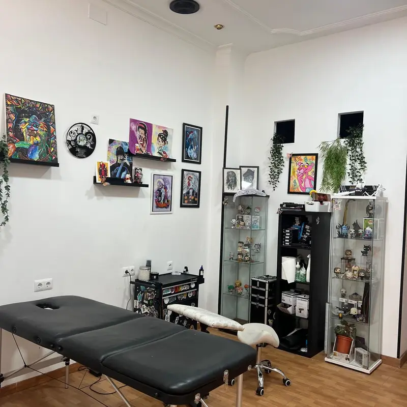 Zona de trabajo del estudio con camilla de tatuaje negra, paredes blancas con cuadros, estanterías y vitrinas con figuras decorativas y plantas.