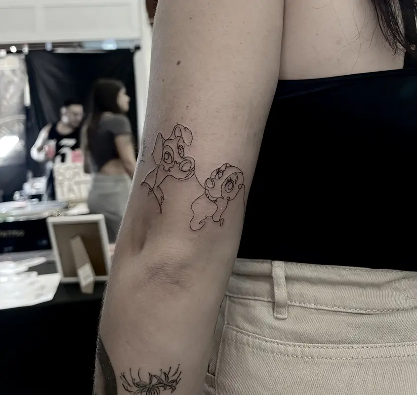 Pareja de personajes dibujados con línea continua, estilo cartoon minimalista en el brazo.