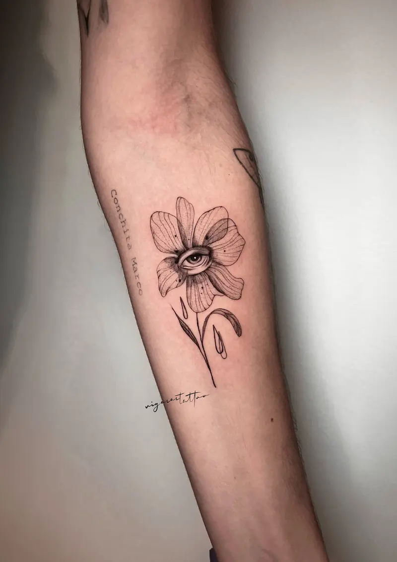 Flor con un ojo en el centro, mezcla de línea fina y sombreado suave; acompañada de nombres en vertical.